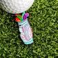 A Bag-O-Dicks Ball Marker by Logie Joe's Golf sits on green turf, shaped like a golf bag with playful stiff shafts, labeled "Happy Pills" in pink script, alongside a golf ball.