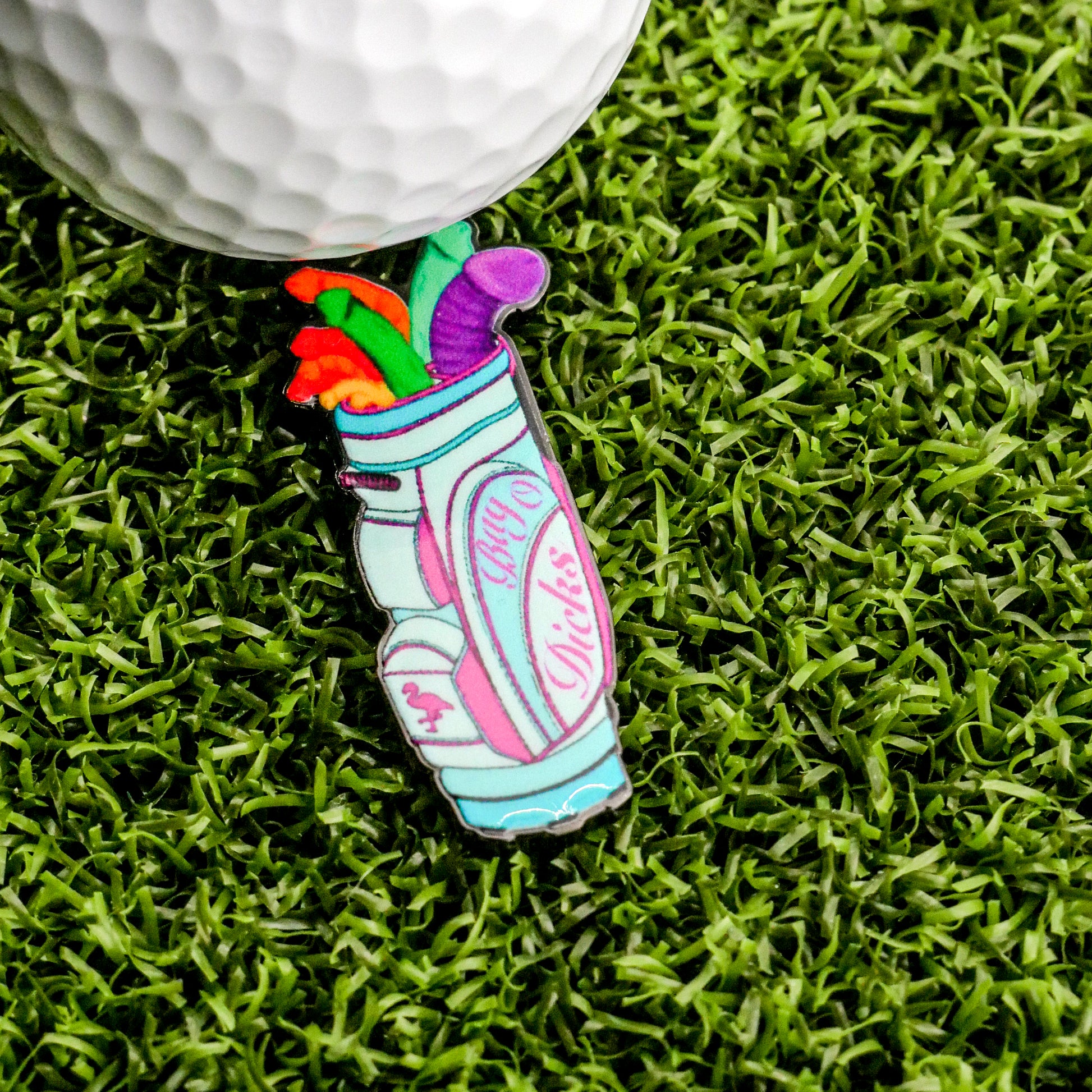 A Bag-O-Dicks Ball Marker by Logie Joe's Golf sits on green turf, shaped like a golf bag with playful stiff shafts, labeled "Happy Pills" in pink script, alongside a golf ball.