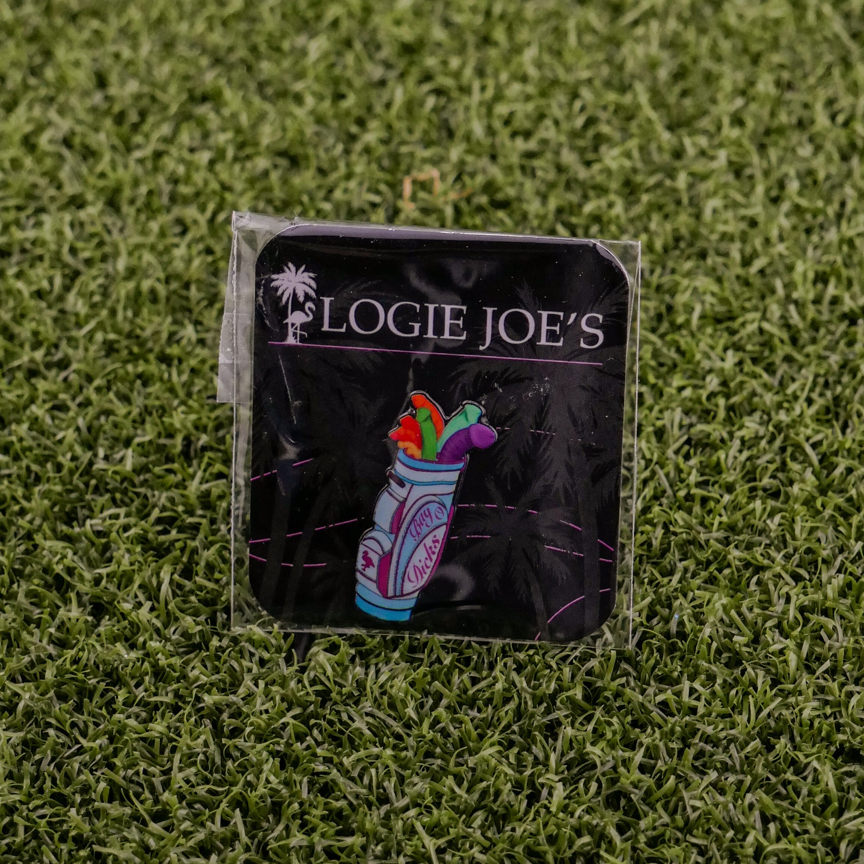 A Bag-O-Dicks pin by Logie Joe's Golf, featuring a vibrant golf bag and clubs design with logo text, is displayed on a black card atop green grass—an ideal accessory for avid golf fans.