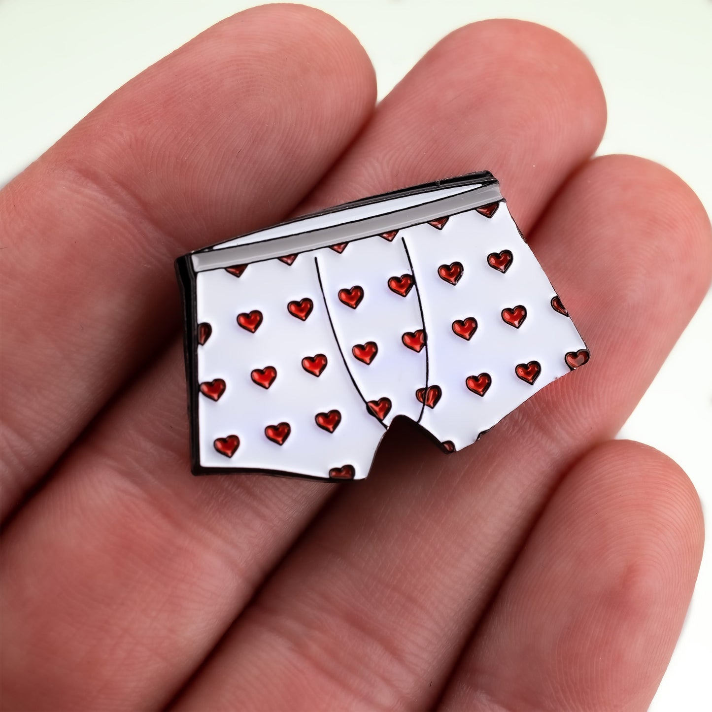 A hand holds the Heart Boxers Ball Marker by Pins and Aces, a magnetic pin shaped like white boxer shorts with a red heart pattern.