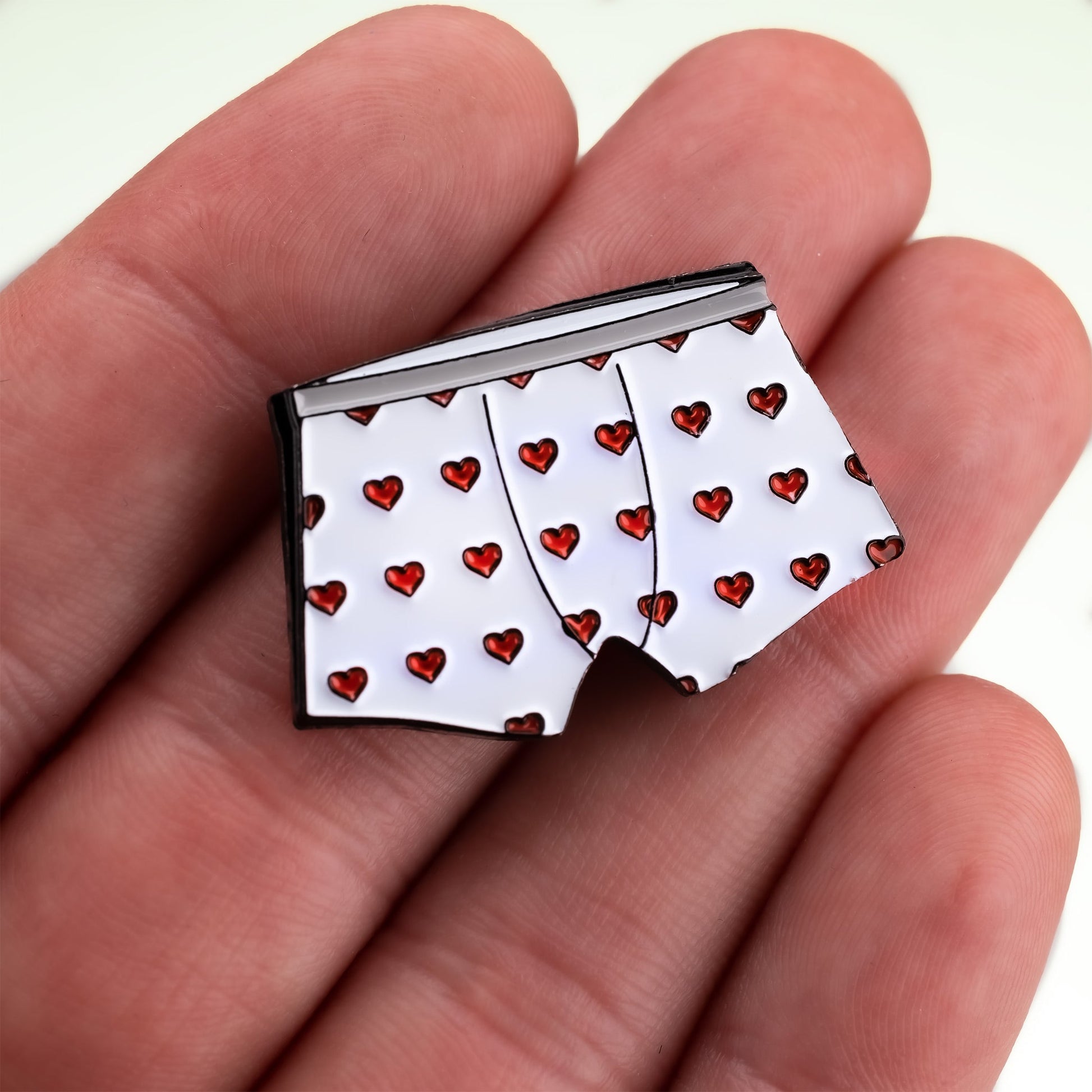 A hand holds the Heart Boxers Ball Marker by Pins and Aces, a magnetic pin shaped like white boxer shorts with a red heart pattern.