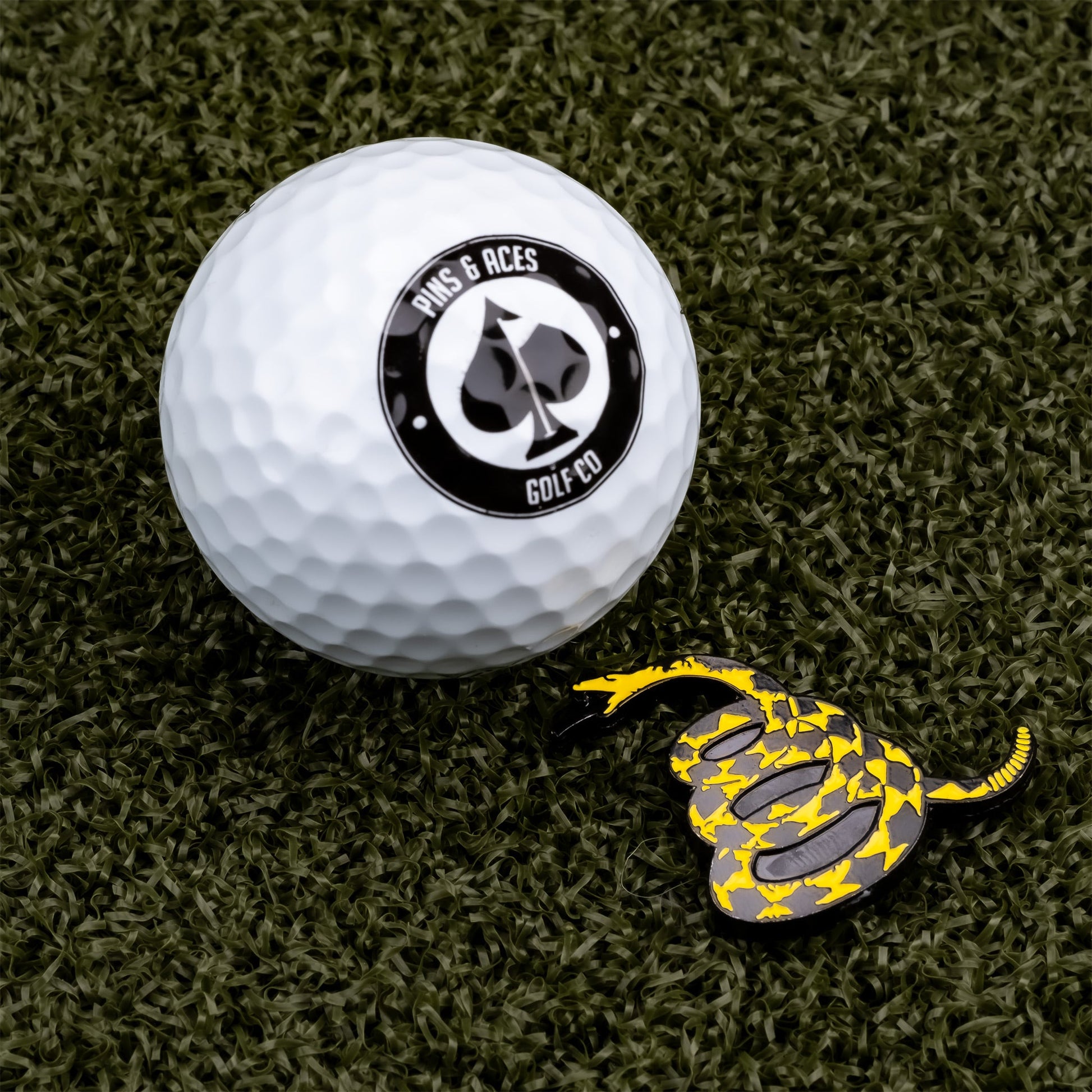 A Pins and Aces golf ball sits on artificial green turf next to the Don't Tread on Me Ball Marker, a yellow and black snake-shaped stainless steel marker.
