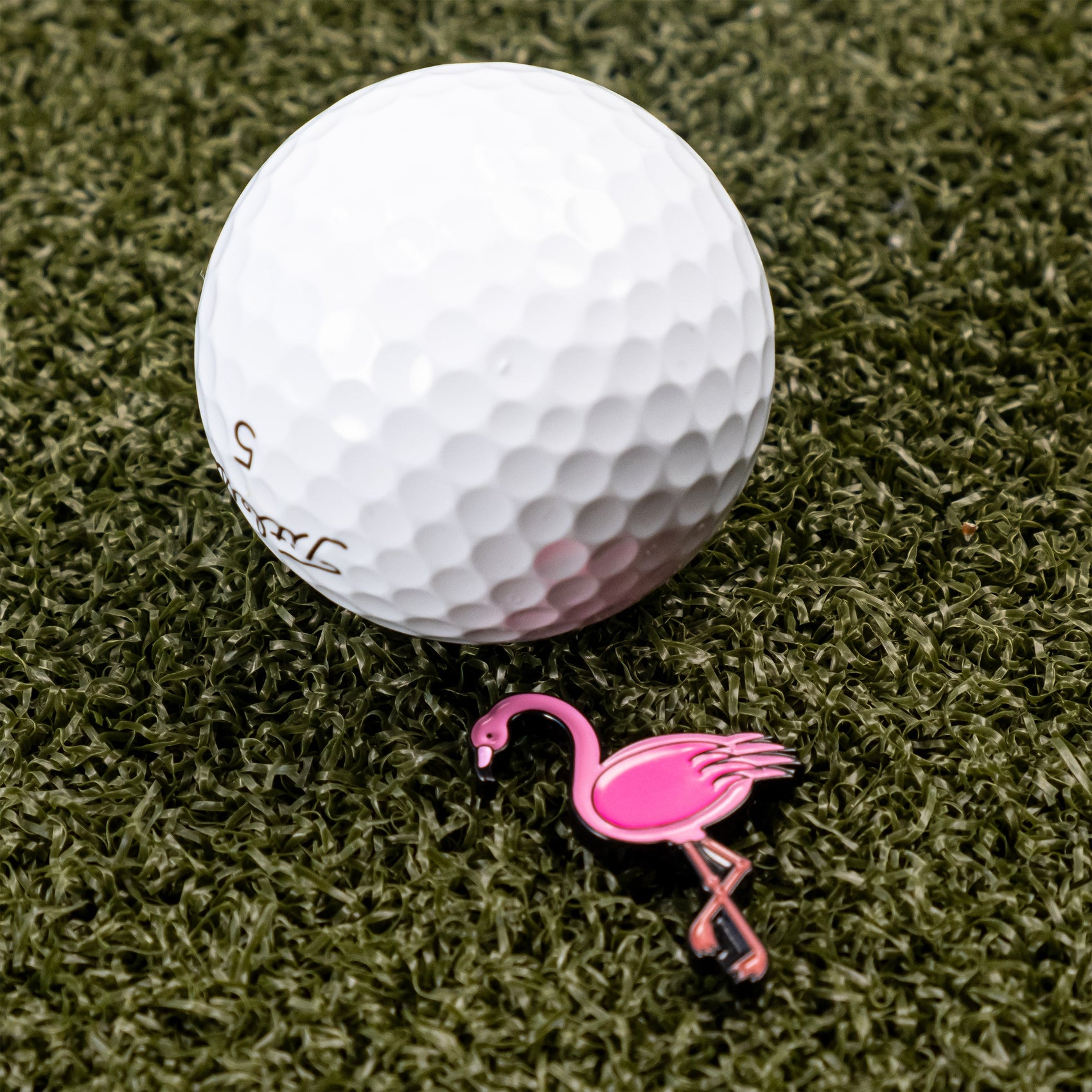 A white golf ball rests on artificial green grass beside the Flamingo Ball Marker by Pins and Aces, adding a cute touch to any round.