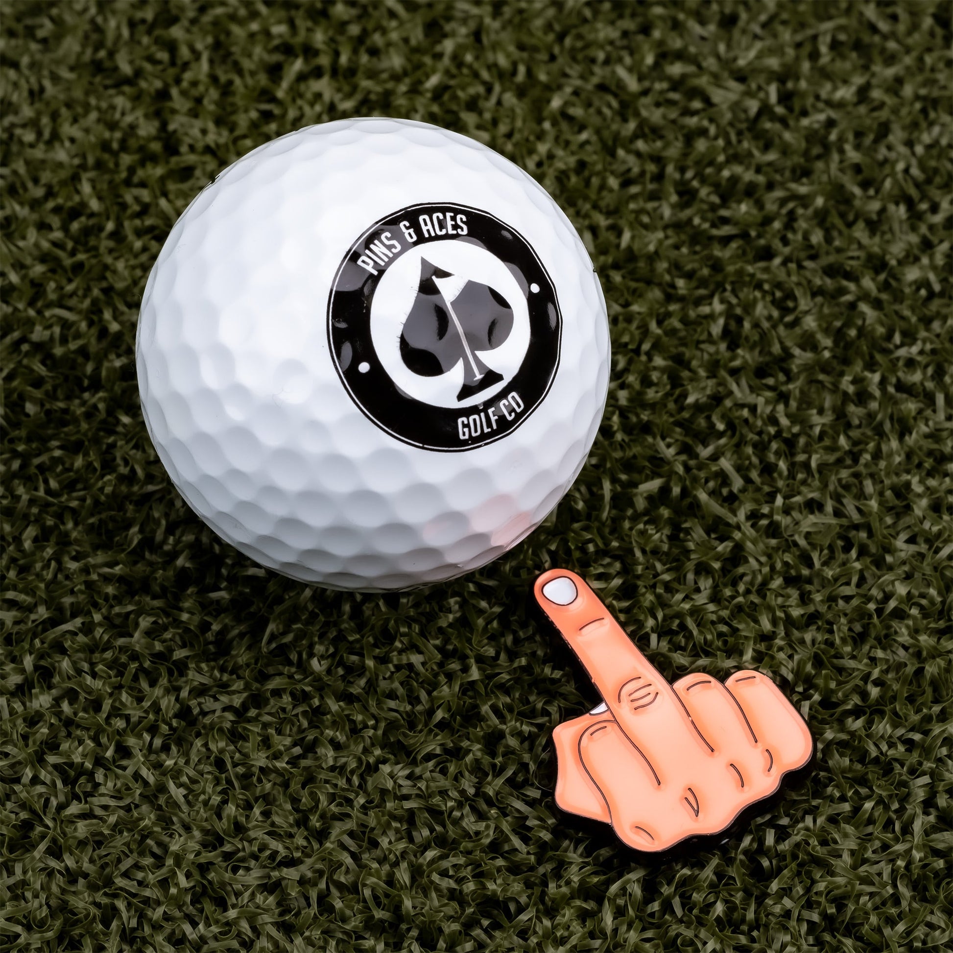 A Pins and Aces golf ball rests on artificial turf next to the Middle Finger Ball Marker—a cartoonish, stainless steel hand with its middle finger raised.