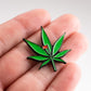 A hand holds the Pins and Aces Pot Leaf Ball Marker—an enamel pin shaped like a green cannabis leaf with a small red flag, featuring a magnetic stainless steel backing, perfect as a unique golf ball marker.