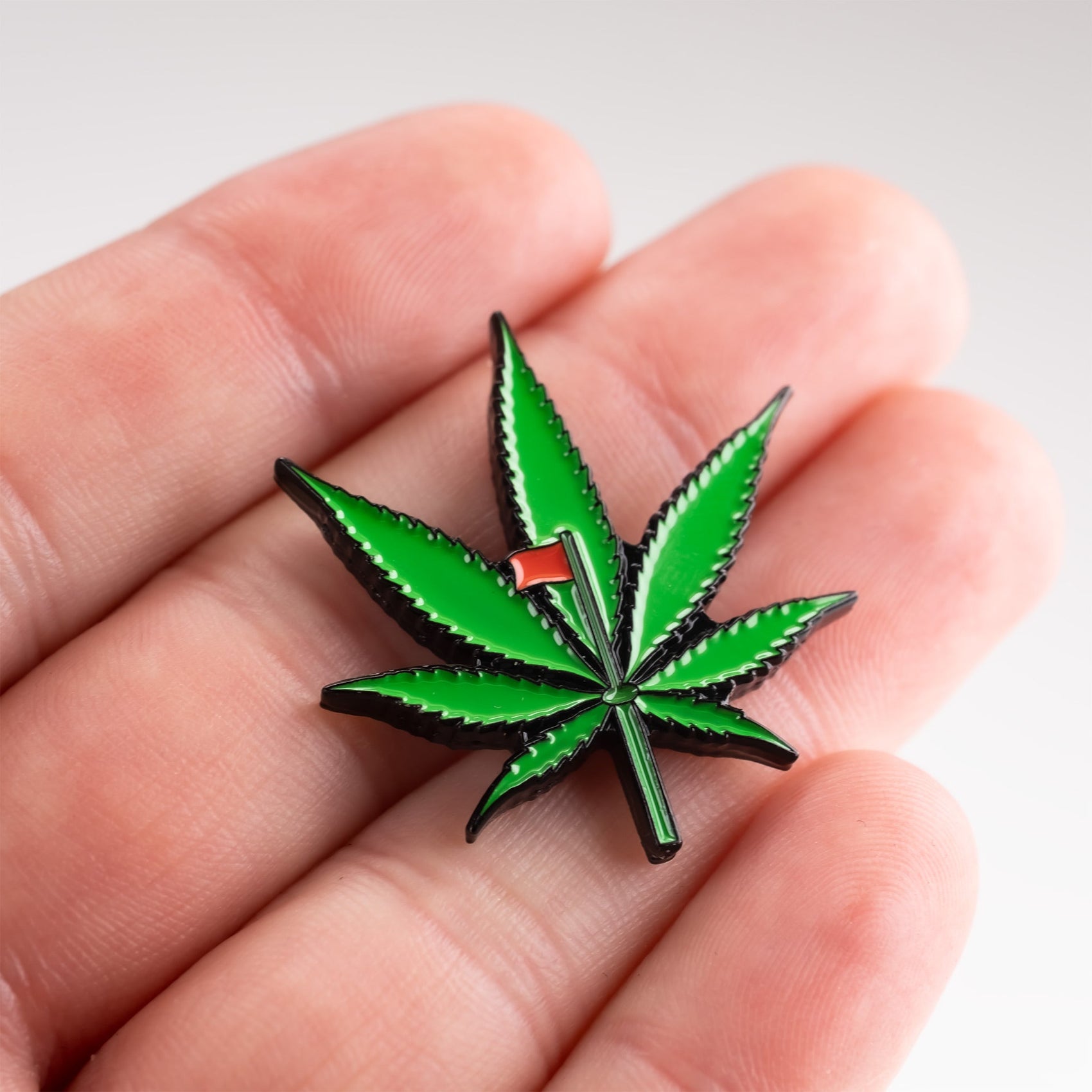 A hand holds the Pins and Aces Pot Leaf Ball Marker—an enamel pin shaped like a green cannabis leaf with a small red flag, featuring a magnetic stainless steel backing, perfect as a unique golf ball marker.