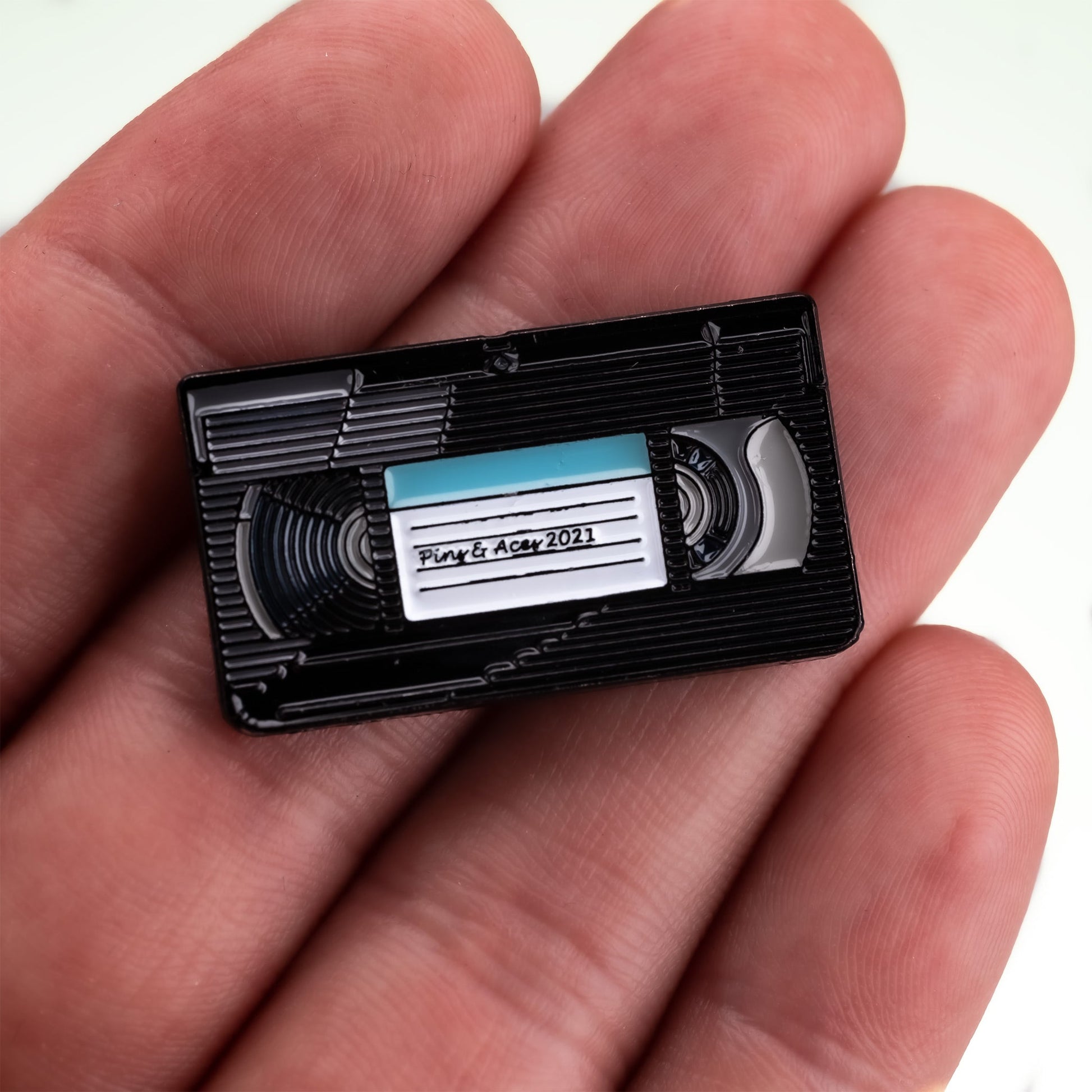 A person holds a small, black VHS Ball Marker by Pins and Aces with a handwritten “Pink & Aqua 2021” label. This detailed marker is about the size of an enamel or magnetic ball marker used on the golf course.