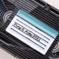 Close-up of a black plastic VHS Ball Marker by Pins and Aces, featuring a white and teal label marked "Pin & Acen 2021" in black cursive—evoking the look of collectible enamel or distinctive golf ball markers.