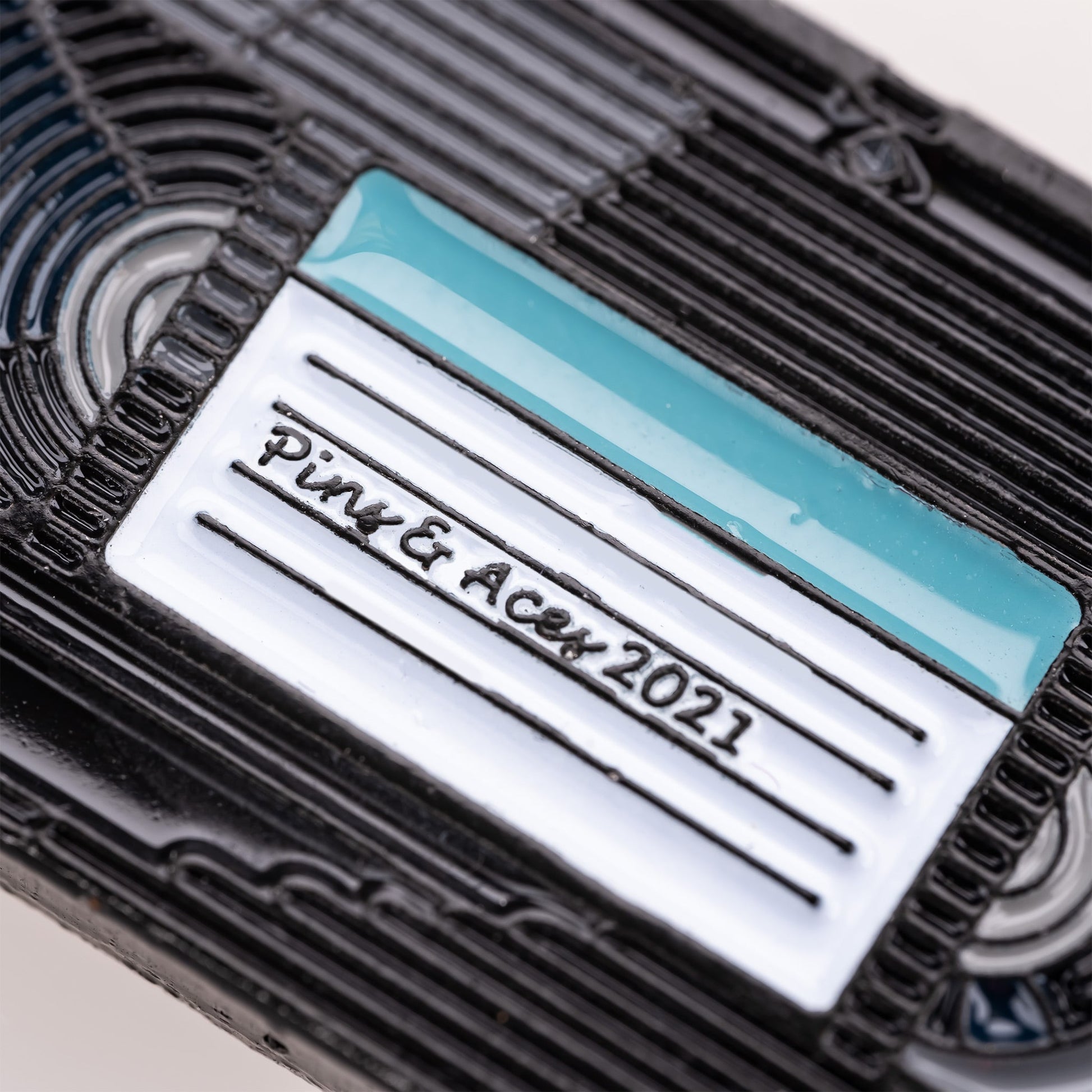 Close-up of a black plastic VHS Ball Marker by Pins and Aces, featuring a white and teal label marked "Pin & Acen 2021" in black cursive—evoking the look of collectible enamel or distinctive golf ball markers.