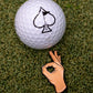 A white golf ball with a black spade symbol rests on green turf beside the Pins and Aces OK Hand Ball Marker, a premium enamel golf accessory shaped like a hand making an OK gesture.
