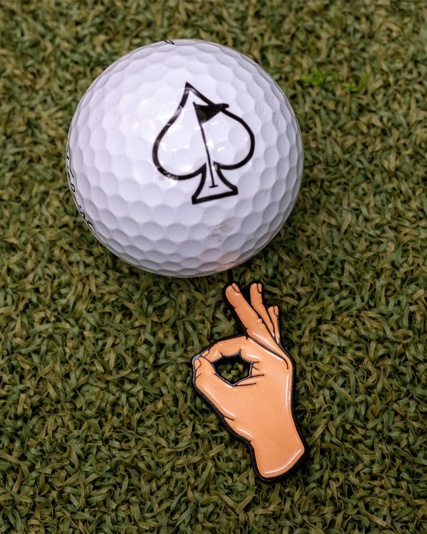 A white golf ball with a black spade symbol rests on green turf beside the Pins and Aces OK Hand Ball Marker, a premium enamel golf accessory shaped like a hand making an OK gesture.
