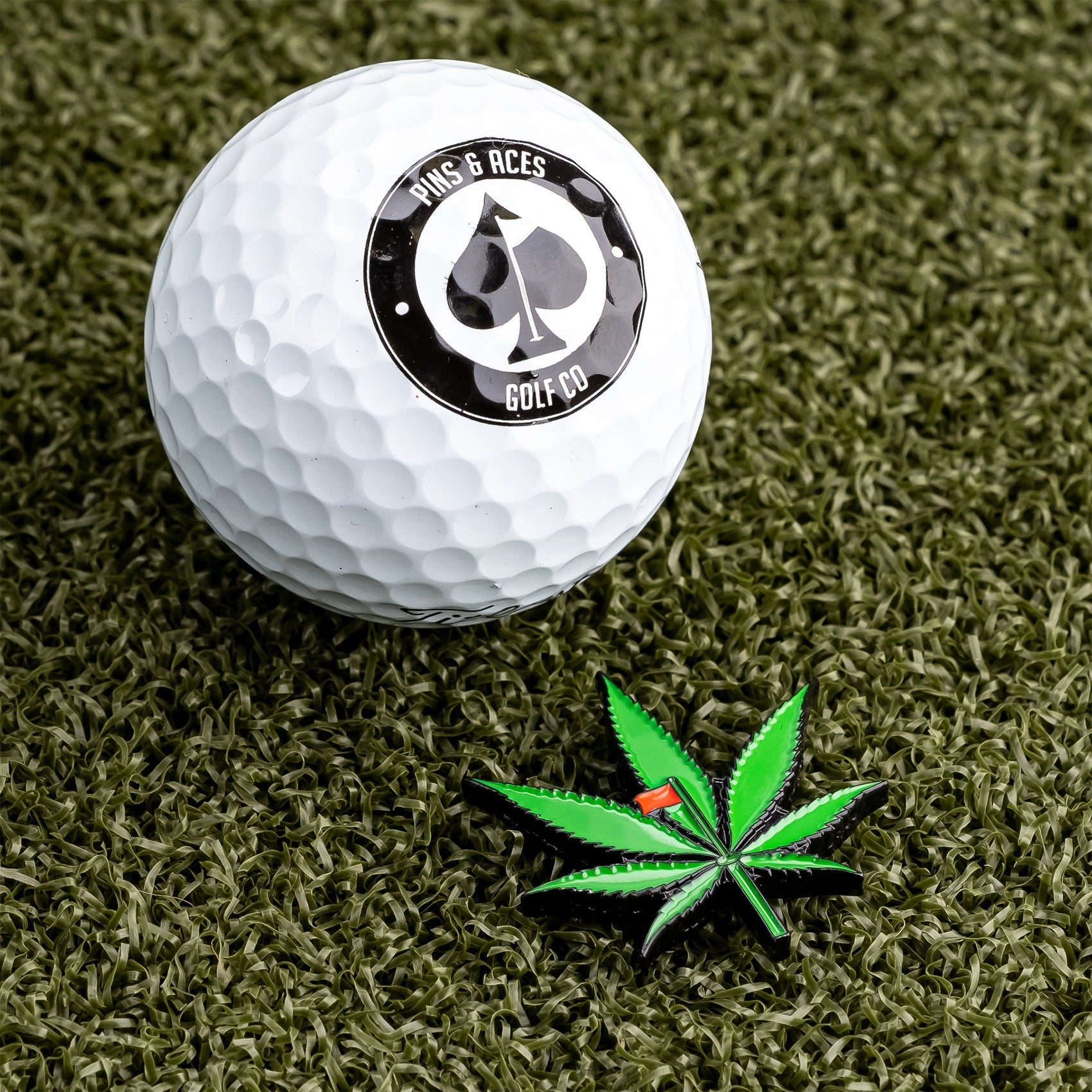 A white golf ball with a Pins and Aces logo rests on green turf beside the Pot Leaf Ball Marker—an enamel pin shaped like a green cannabis leaf with a red tip, perfect as a unique golf ball marker.