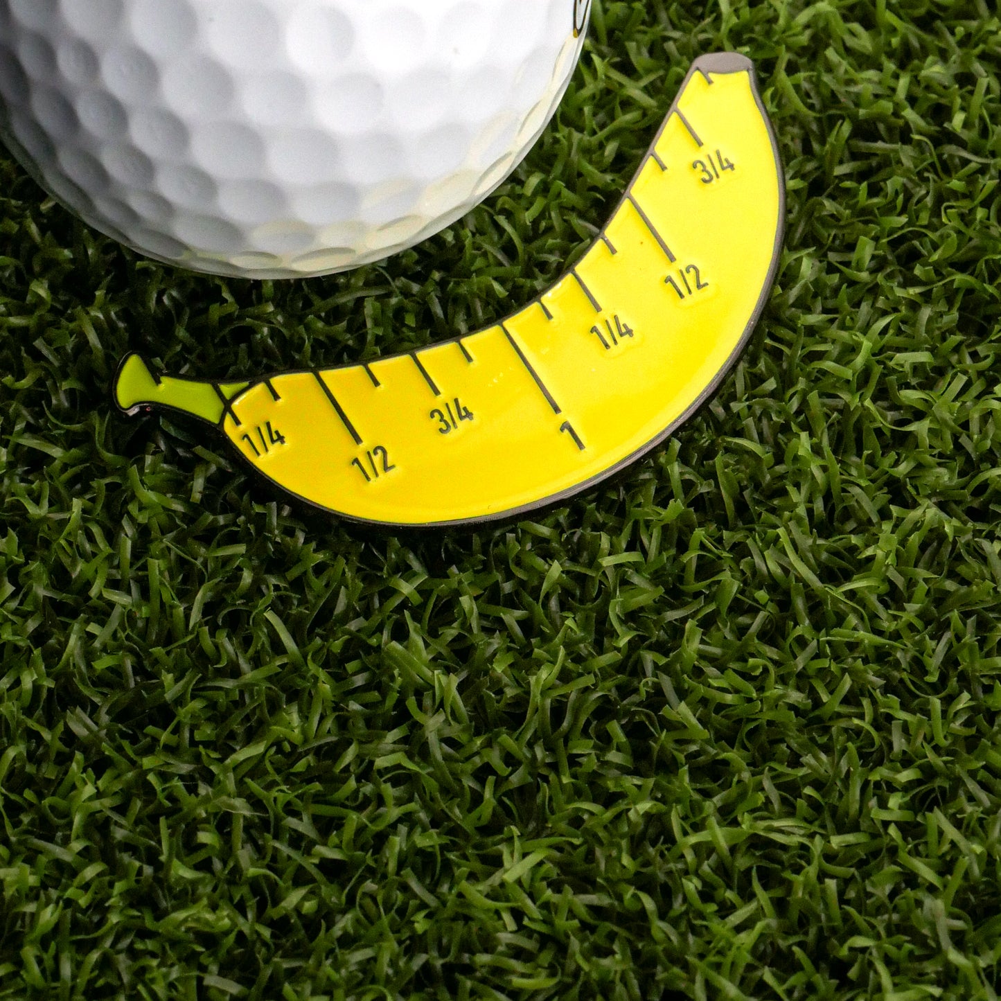 A close-up of Logie Joe's Golf Banana For Scale Ball Marker—a yellow, banana-shaped tool marked from 1/4 to 1 1/2 units—next to a golf ball on artificial grass.