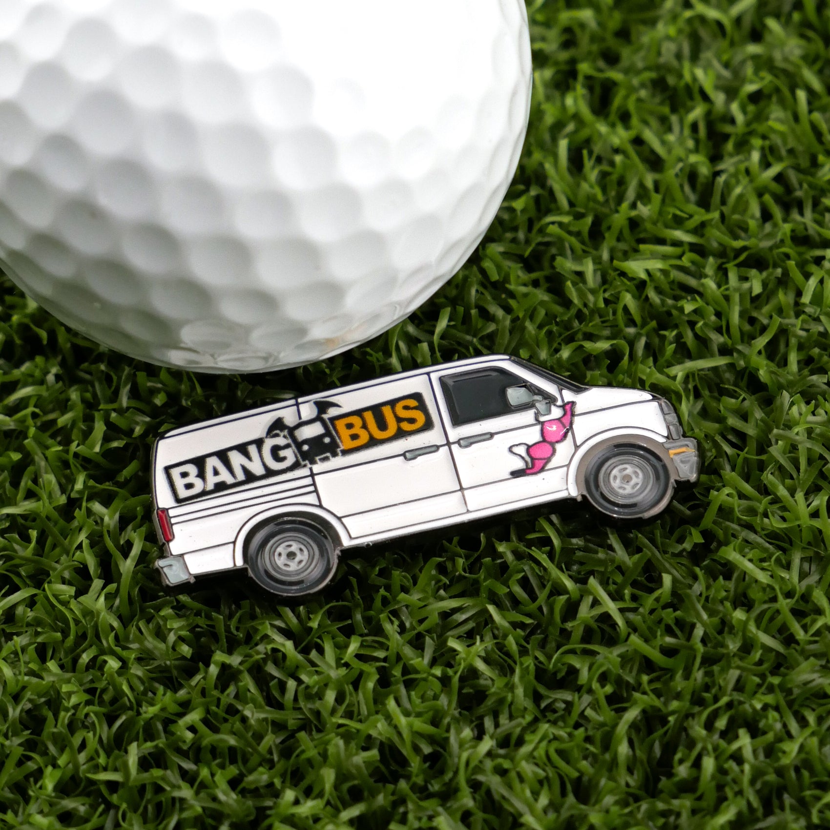 A unique golf accessory, the Bang Bus by Logie Joe's Golf is a white van-shaped ball marker with a pink cartoon figure and "BANG BUS" label, shown on artificial green grass beside a white golf ball.