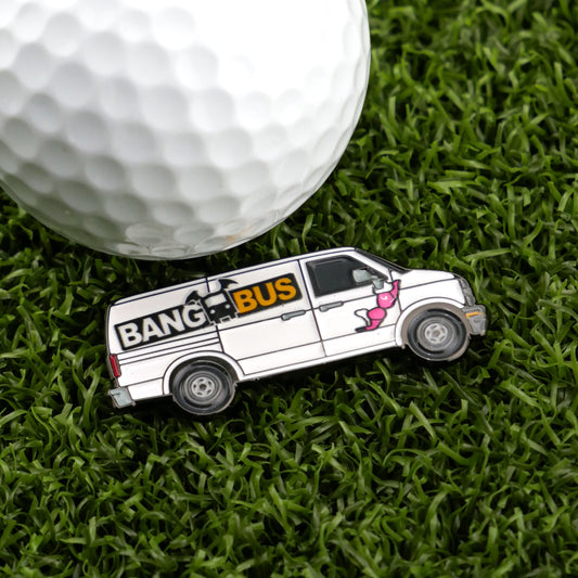 A golf ball rests on artificial grass beside Logie Joe's Golf Bang Bus Ball Marker, a white van-shaped pin featuring “BANG BUS” text and a pink-clad cartoon woman—one of the most unique premium golf accessories.