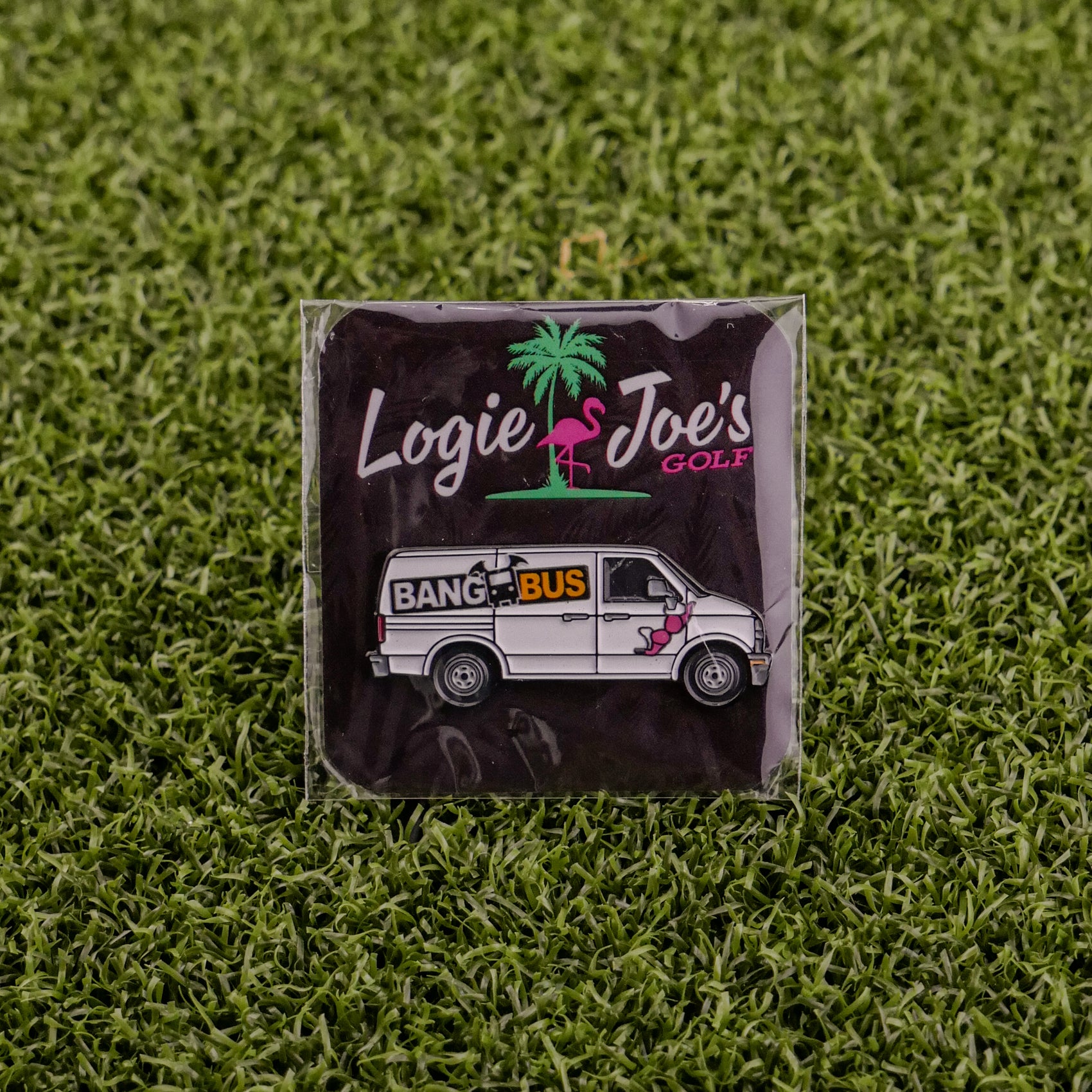 A square pin named Bang Bus from Logie Joe’s Golf features a van with a flamingo, displayed on green artificial grass and packaged on a palm tree card—one of our unique golf accessories.