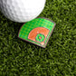 The Ballmarkers.com Baseball Field Ball Marker, shaped like a baseball infield, and a golf ball rest on green artificial grass, creating a unique golf putting aid.