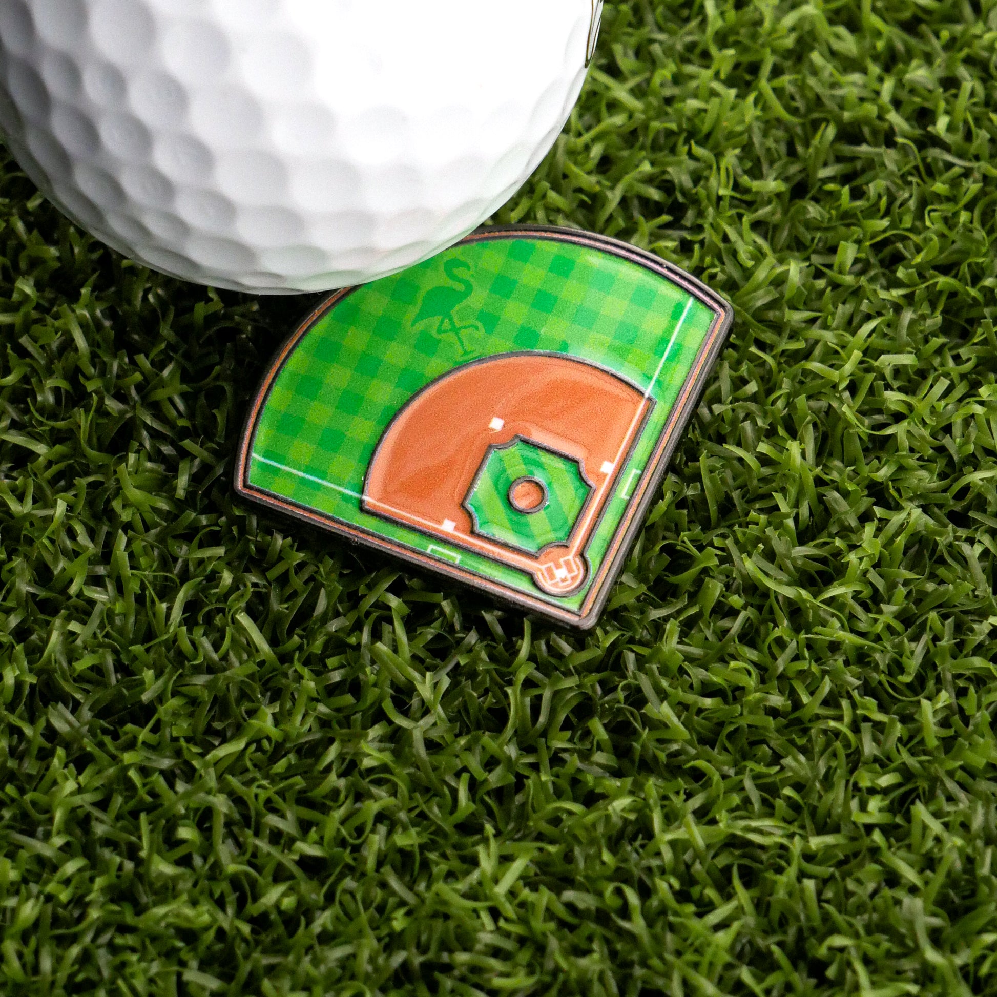 The Ballmarkers.com Baseball Field Ball Marker, shaped like a baseball infield, and a golf ball rest on green artificial grass, creating a unique golf putting aid.