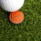 A white golf ball and a Ballmarkers.com Basketball Ball Marker, featuring a basketball pattern, rest on green artificial grass—ideal for working on your short game.
