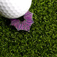 A white golf ball sits on artificial green grass next to the Bat Ball Marker by Ballmarkers.com—a metallic marker shaped like a bat featuring a striking purple and black geometric design.