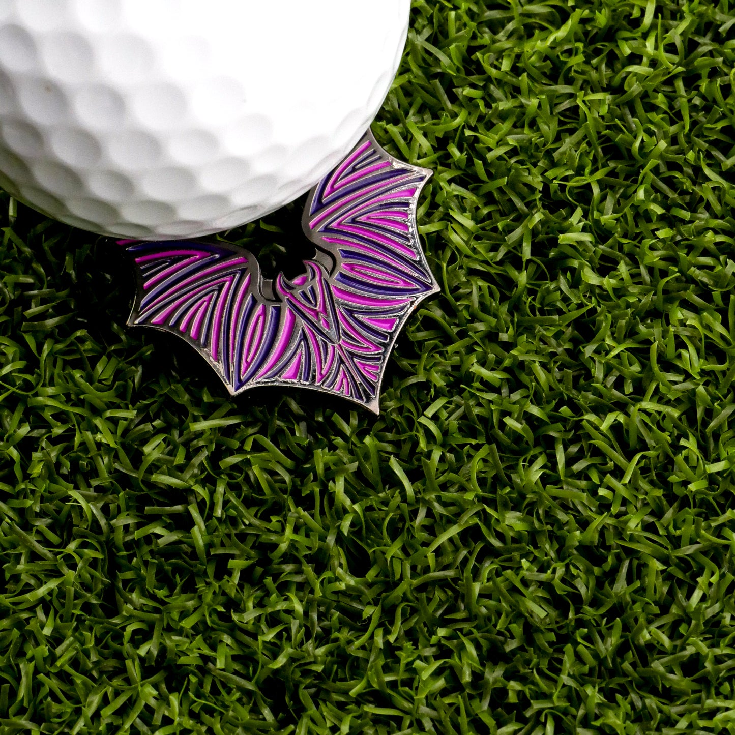 A white golf ball sits on artificial green grass next to the Bat Ball Marker by Ballmarkers.com—a metallic marker shaped like a bat featuring a striking purple and black geometric design.