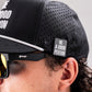 Close-up of a person wearing a black perforated cap with a Pins and Aces "Be A Good Person" Ball Marker attached, plus black goodr sunglasses. Their curly hair and only part of their face are visible.