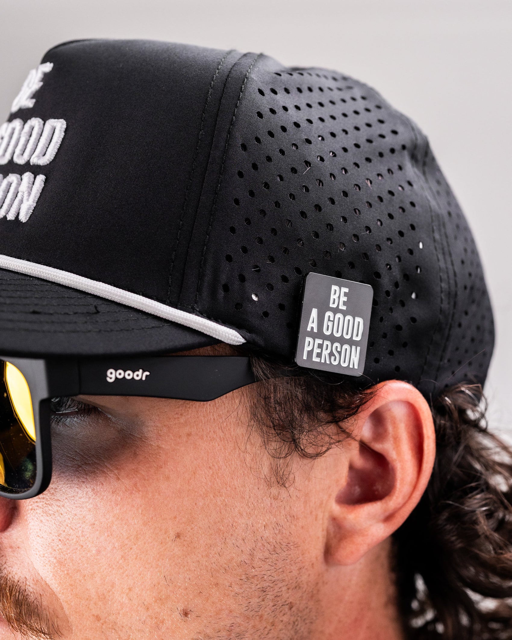 Close-up of a person wearing a black perforated cap with a Pins and Aces "Be A Good Person" Ball Marker attached, plus black goodr sunglasses. Their curly hair and only part of their face are visible.