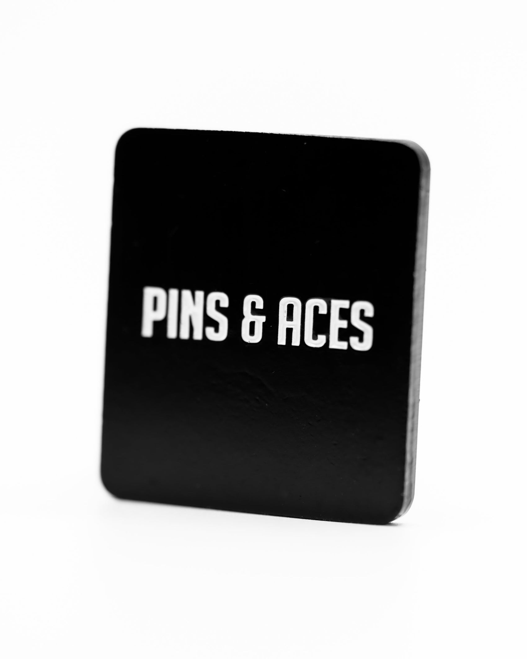 A black rectangular holder with "PINS & ACES" in bold white on the front, made for the Pins and Aces Be A Good Person Ball Marker, stands out against a plain white background.