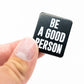 A hand displays the Be A Good Person Ball Marker by Pins and Aces—a sleek black square accessory with white text, perfect for fans of stylish golf gear—set against a plain white background.