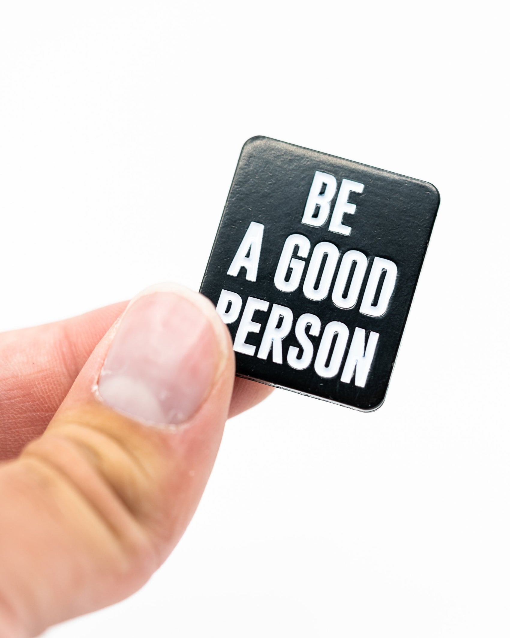 A hand displays the Be A Good Person Ball Marker by Pins and Aces—a sleek black square accessory with white text, perfect for fans of stylish golf gear—set against a plain white background.