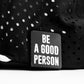 The Pins and Aces Be A Good Person Ball Marker, featuring bold white text on black, is displayed on a perforated fabric background—perfect for golfers who appreciate standout accessories.