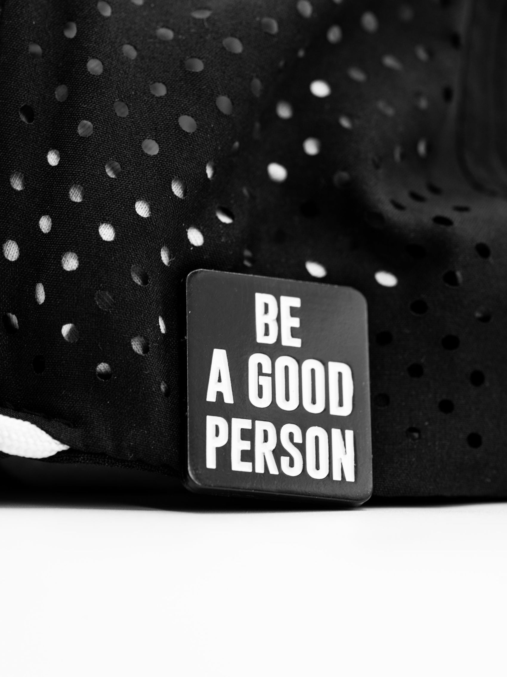 The Pins and Aces Be A Good Person Ball Marker, featuring bold white text on black, is displayed on a perforated fabric background—perfect for golfers who appreciate standout accessories.