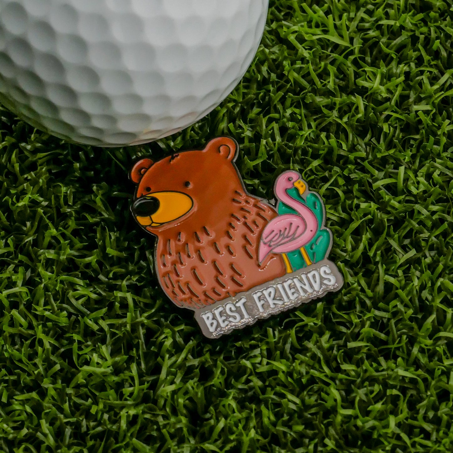 The Best Friends Ball Marker by Logie Joe's Golf is a colorful, premium pin with a brown bear, pink flamingo, and "BEST FRIENDS" text, sitting on green artificial grass next to a golf ball—an ideal fun golf ball marker.