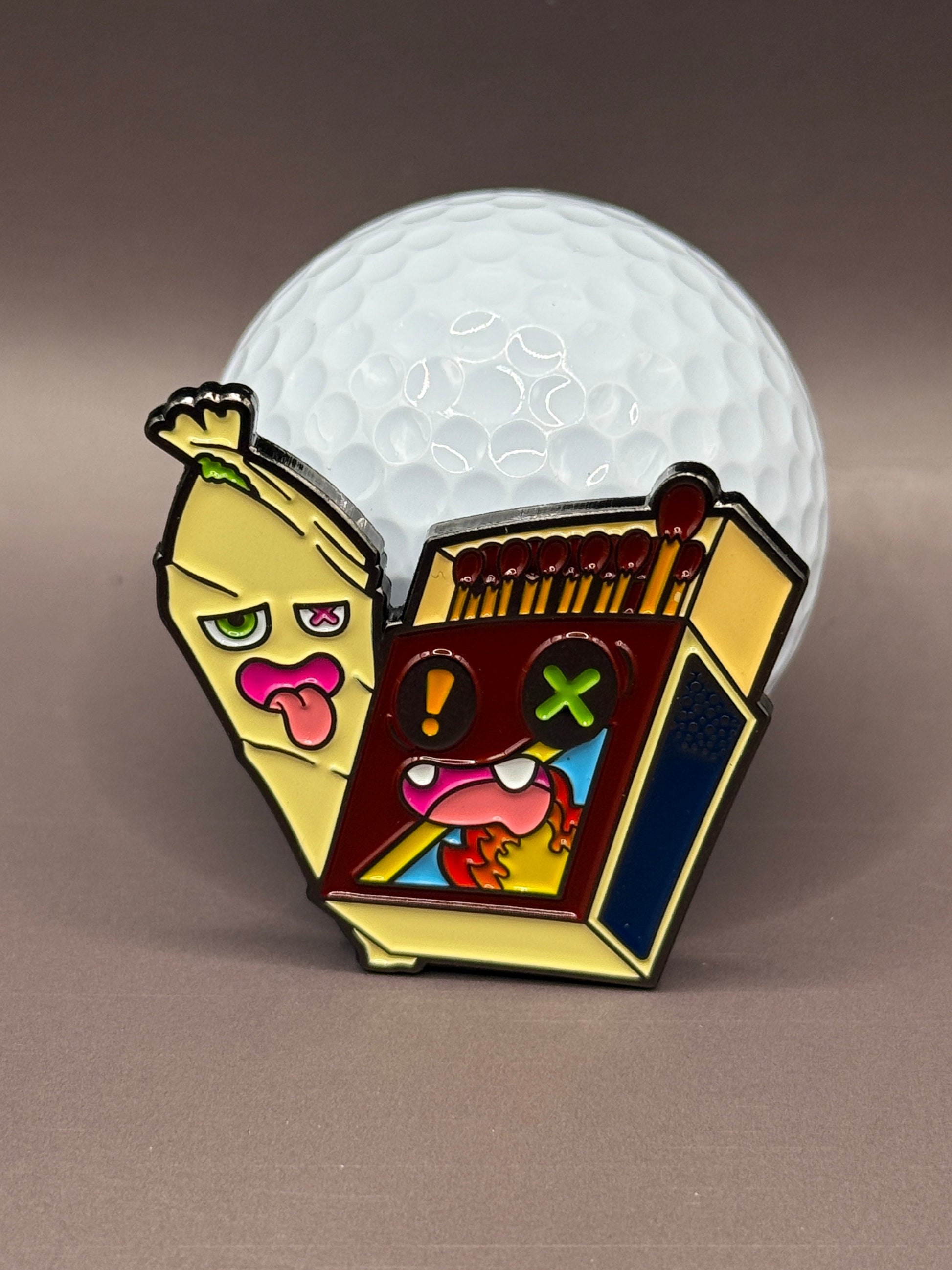 The Best Buds Ball Marker by Crown Golf Designs is a vibrant metal enamel pin set, featuring a quirky banana and matchbox duo with expressive faces, displayed in front of a golf ball on a neutral background.