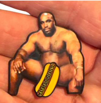 Golf Gear Heaven’s The Big Glizzy Ball Marker is a cheeky sticker featuring a muscular, nude, bald man seated with a cartoon hot dog covering his crotch, shown being held between someone’s fingers.