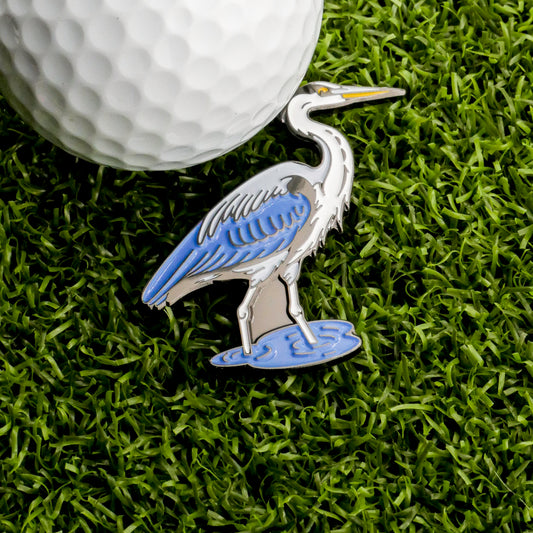 A Great Blue Heron Ball Marker by Ballmarkers.com, featuring a blue and white heron design, rests on green grass beside a golf ball—an ideal nature-inspired accessory for golfers seeking a unique ball marker.