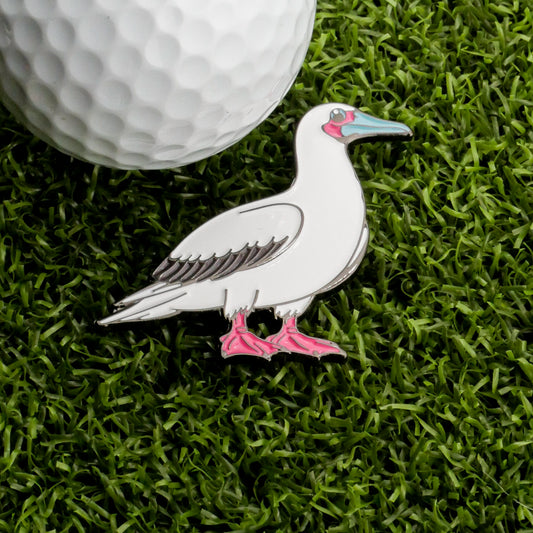 The Red-Footed Booby Ball Marker by Ballmarkers.com features a white bird pin with pink feet and a blue beak, perched on green grass next to a white golf ball—making it a charming and unique addition to your golf accessories collection.