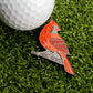 A metal Cardinal Ball Marker from Ballmarkers.com, designed in the shape of a red cardinal, rests on artificial grass beside a white golf ball—an ideal wildlife-themed accessory for any golf enthusiast.