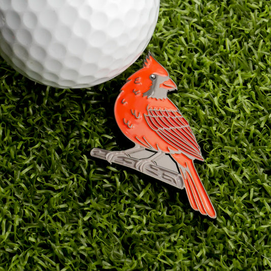 A metal Cardinal Ball Marker from Ballmarkers.com, designed in the shape of a red cardinal, rests on artificial grass beside a white golf ball—an ideal wildlife-themed accessory for any golf enthusiast.