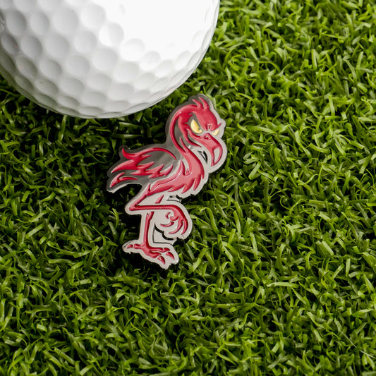 A golf ball sits on artificial grass next to the Jogie Loes Evil Flamingo Ball Marker by Ballmarkers.com, which features a cartoon flamingo with an intense expression and is custom cut to shape.