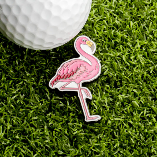A golf ball rests beside the Ballmarkers.com Flamingo Ball Marker on vibrant green artificial grass, highlighting this playful accessory that brings tropical flair to your game.