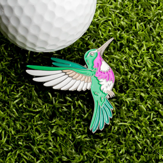A charming Hummingbird Ball Marker from Ballmarkers.com, featuring green, pink, and white details, rests on green grass next to a white golf ball—perfect for golfers seeking unique accessories.
