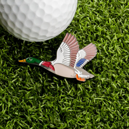 A Mallard Ball Marker by Ballmarkers.com, shaped like a flying duck and crafted in enamel, rests on green grass beside a white golf ball—an ideal wildlife accessory or stylish golf ball marker.