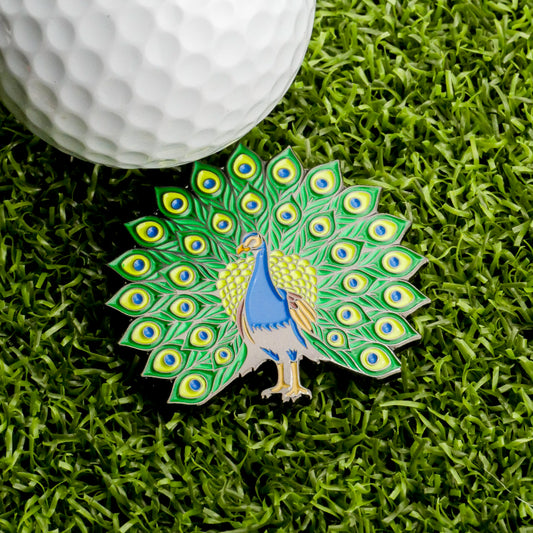 A vibrant Peacock Ball Marker from Ballmarkers.com, featuring a peacock with spread feathers, rests on green grass beside a white golf ball—bringing an exotic and colorful flair to your accessory collection.