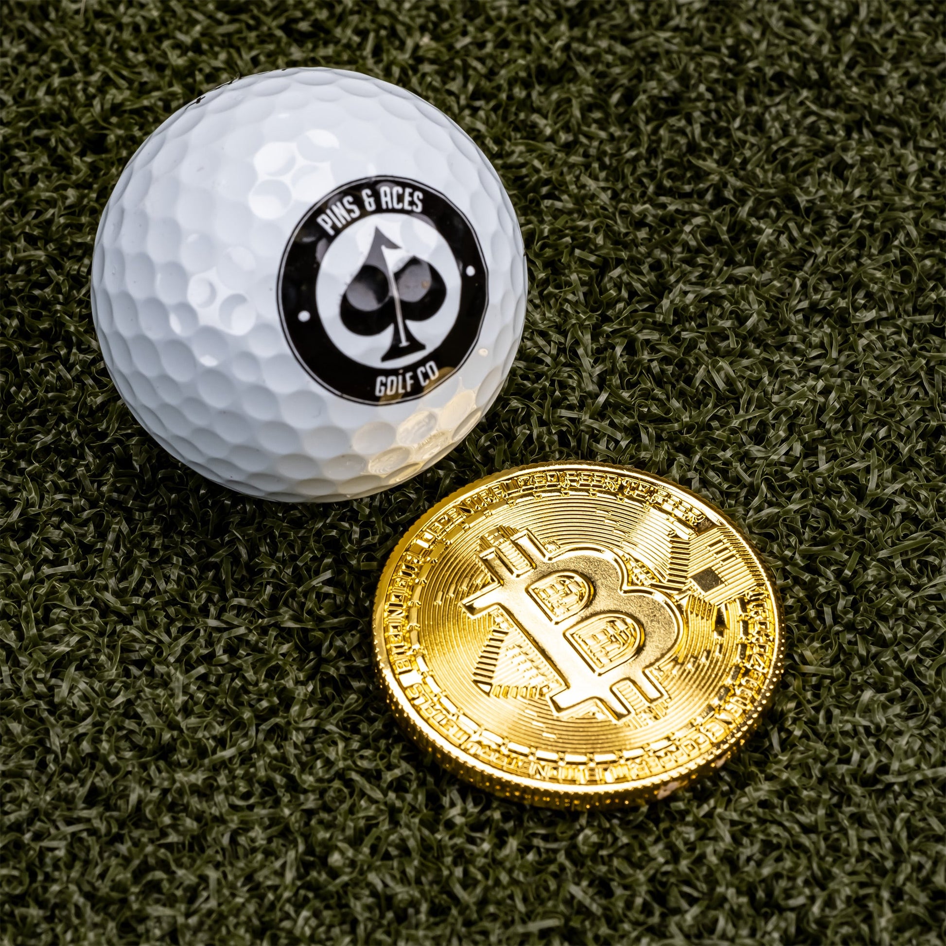 A Pins and Aces white golf ball sits next to the Bitcoin Ball Marker, a shiny gold coin with a Bitcoin symbol and gold chrome finish, both placed on green artificial turf.