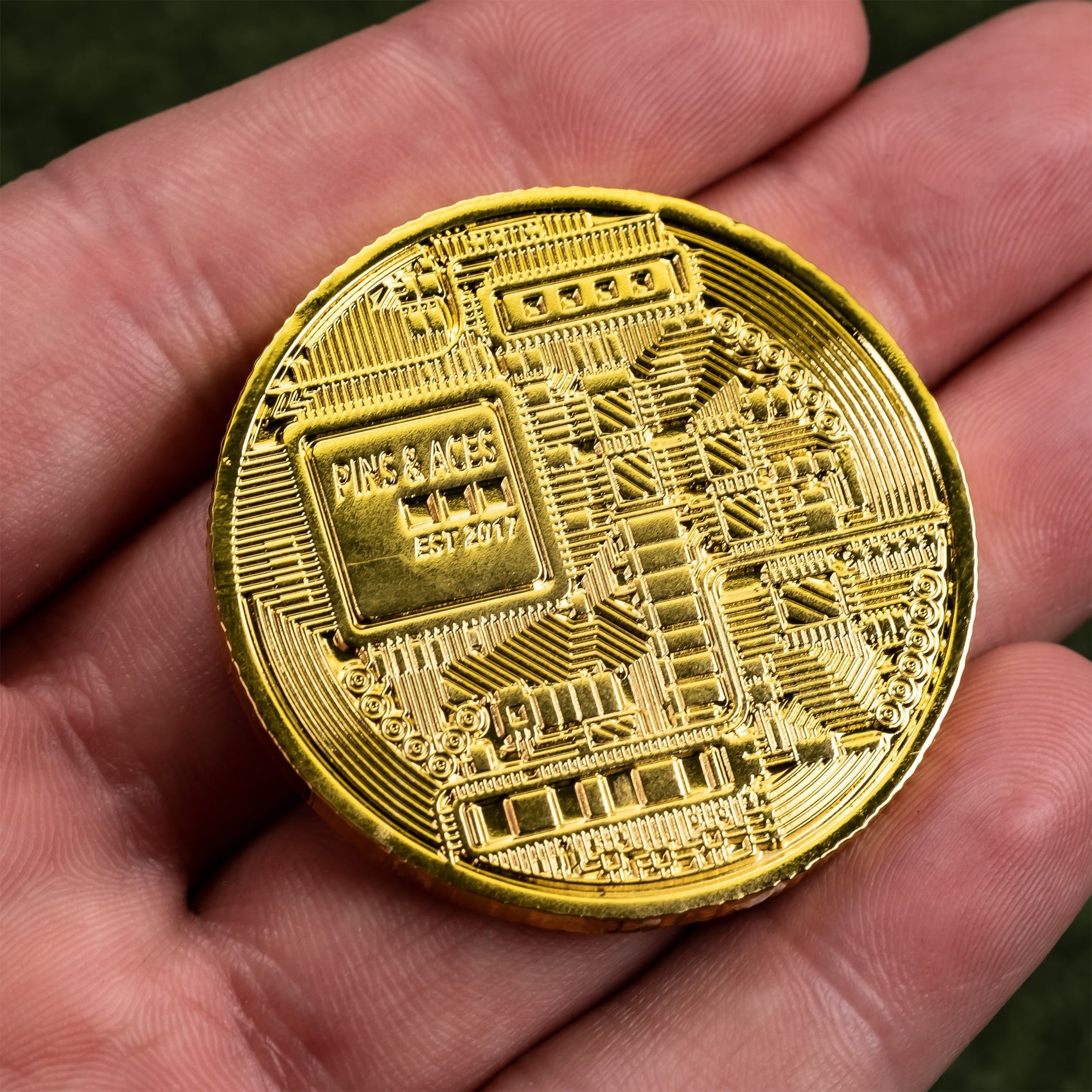 A hand holds the Pins and Aces Bitcoin Ball Marker, featuring a gold chrome finish with intricate circuit-like patterns and "PING & ACES EST. 2017" engraved, inspired by high-end golf ball markers.