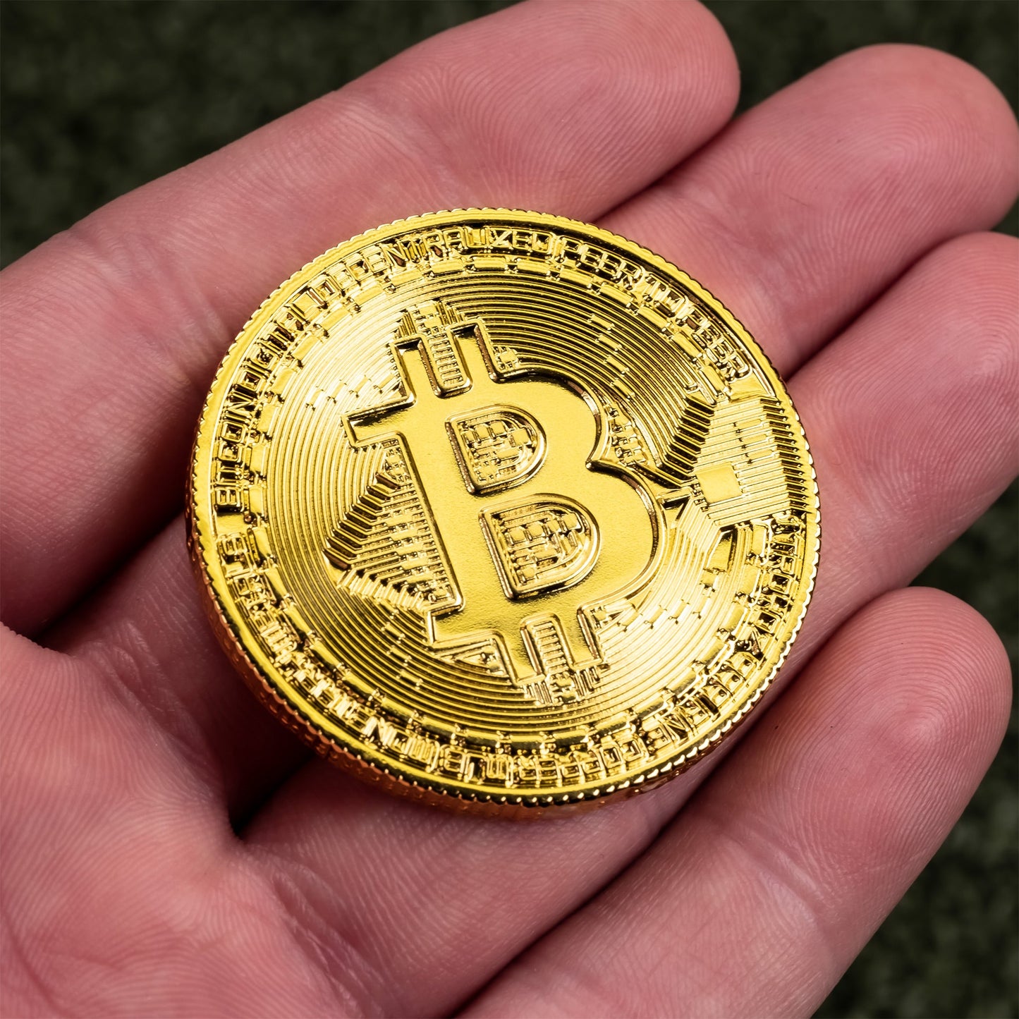 A person holding a shiny gold-colored Bitcoin Ball Marker by Pins and Aces, resembling gold chrome finish golf ball markers, against a blurred green background.