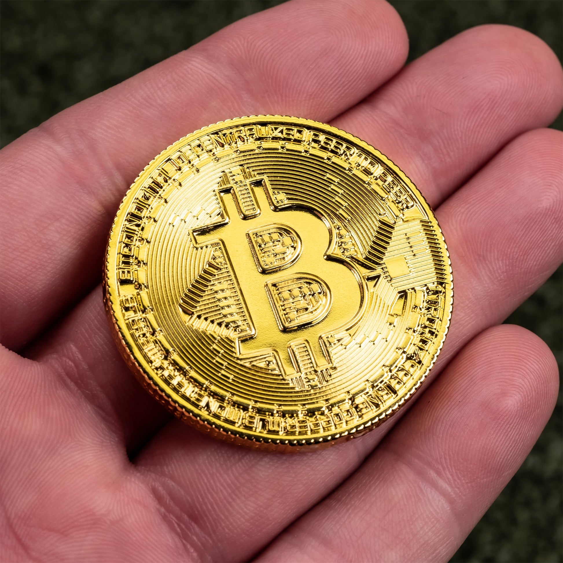 A person holding a shiny gold-colored Bitcoin Ball Marker by Pins and Aces, resembling gold chrome finish golf ball markers, against a blurred green background.