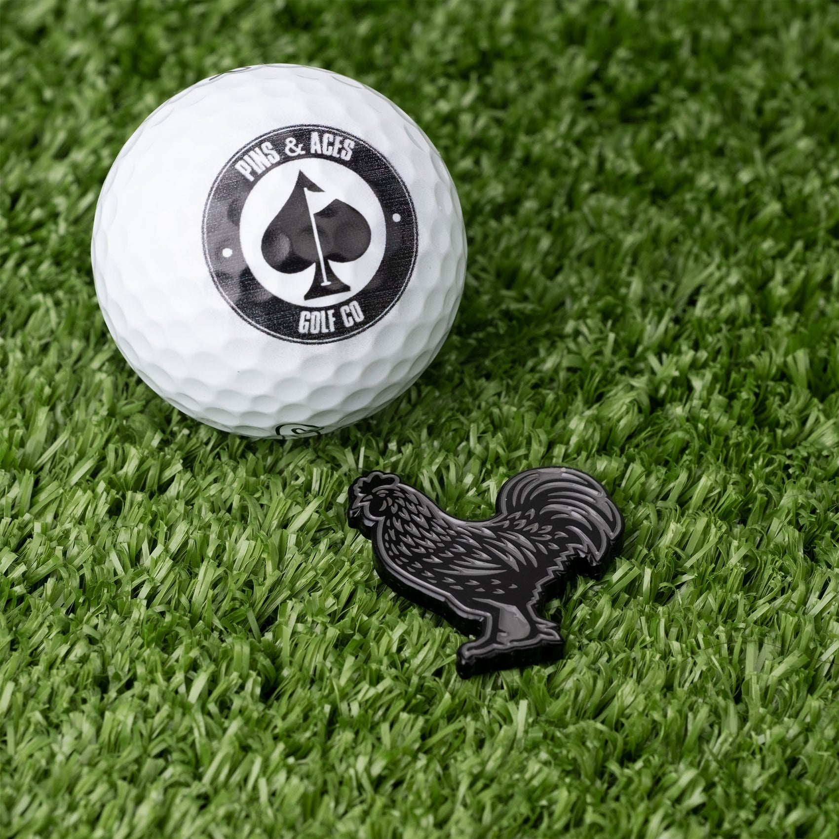 A Pins and Aces golf ball is shown next to the Black Cock Ball Marker—a black enamel rooster-shaped marker—both resting on green artificial turf.