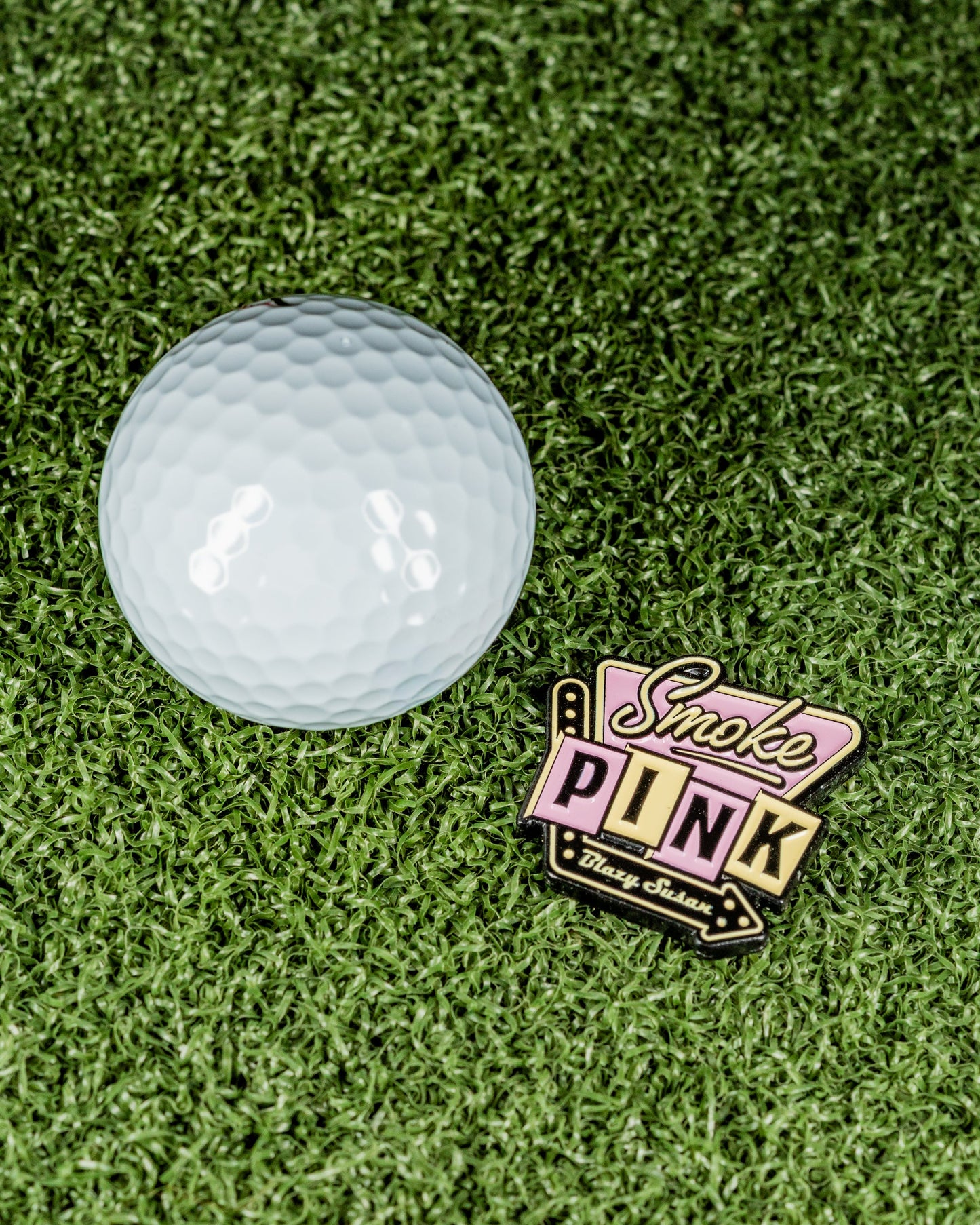 The Pins and Aces Blazy Susan Smoke Pink Ball Marker is a vibrant stainless steel accessory featuring "Smoke PINK, Think Urban," displayed alongside a white golf ball on bright green artificial grass.